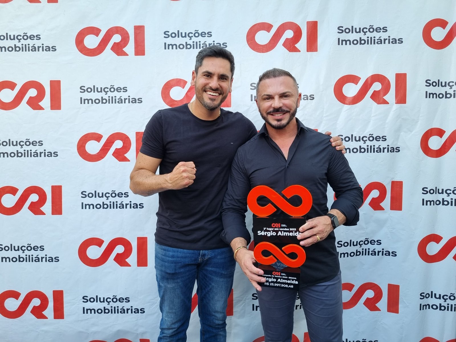 Sérgio Almeida ao lado de um colega, segurando um troféu, diante de painel com o logotipo da CRI